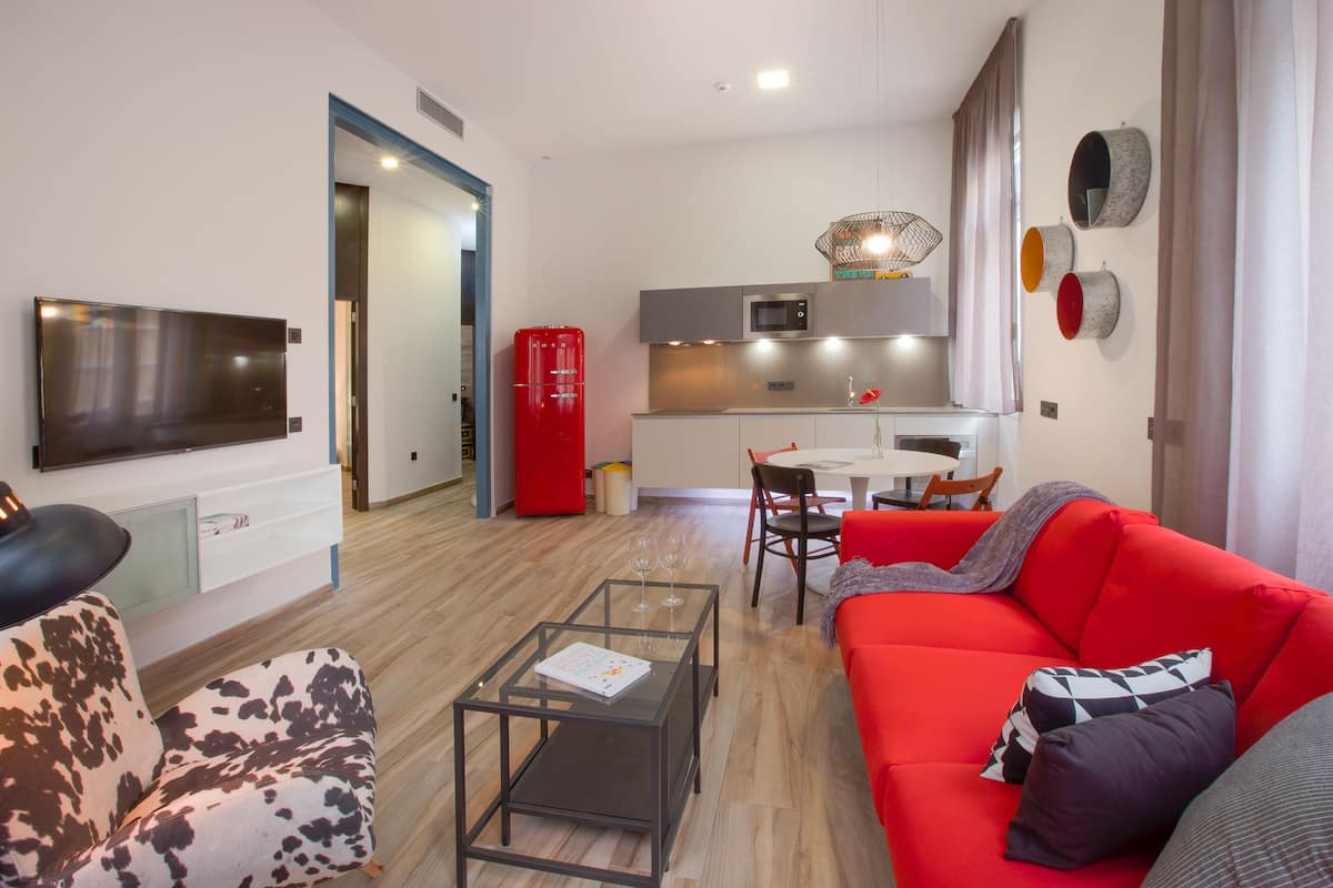 A modern living room with a red sofa, a glass coffee table, and a TV. There's a kitchen area with a red fridge and a dining table.