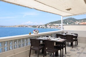 An outdoor terrace with dining tables and chairs, overlooking a coastal town and the sea. - Hotel Karacam (Foça)