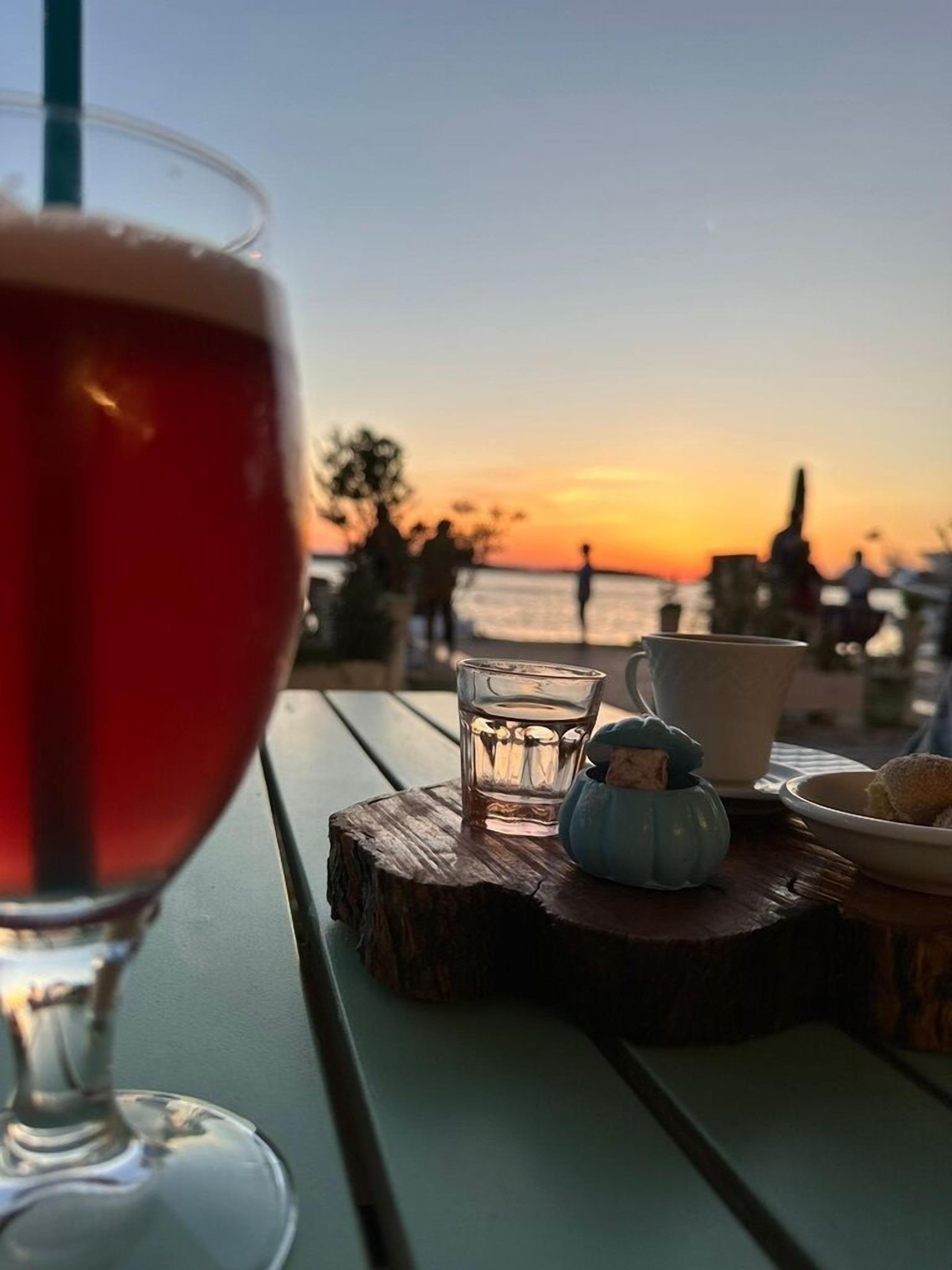 Uma mesa com um copo de bebida vermelha, um copo de água, uma tigela azul pequena e uma xícara branca, com vista para o pôr do sol.