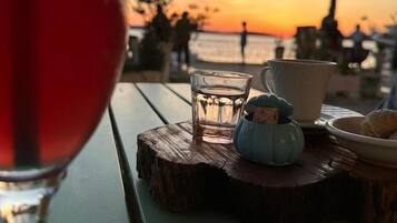 Uma mesa com um copo de bebida vermelha, um copo de água, uma tigela azul pequena e uma xícara branca, com vista para o pôr do sol.