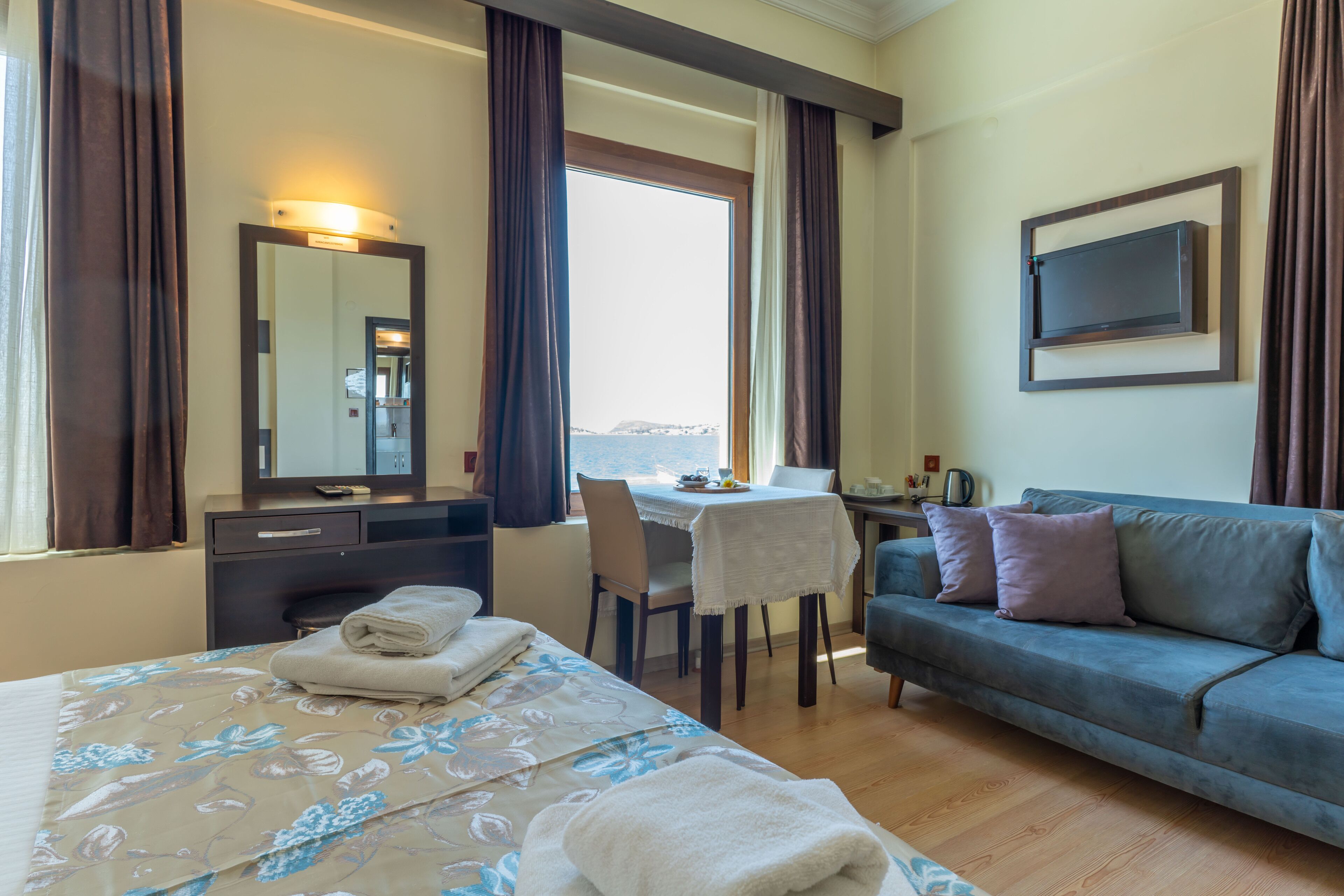 A hotel room with a bed, a sofa, a desk, and a chair. There is a window with curtains, a mirror, and a flat-screen TV mounted on the wall.