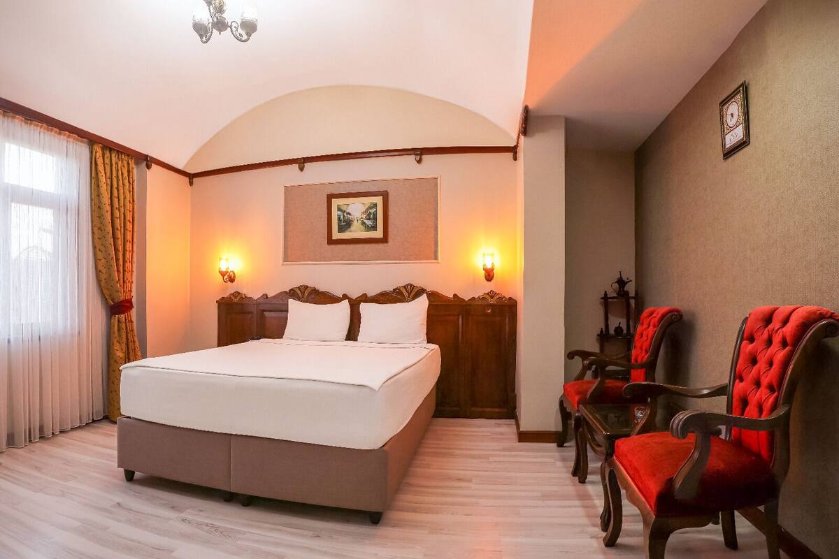 A hotel room with a large bed, two wall-mounted lamps, a framed picture, and two upholstered chairs.