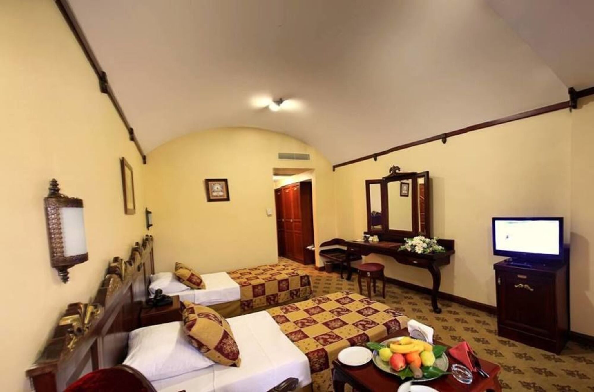 A hotel room with two beds, a desk, a television, and a dining table with fruit.