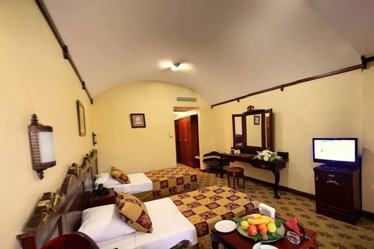 A hotel room with two beds, a desk, a television, and a dining table with fruit.