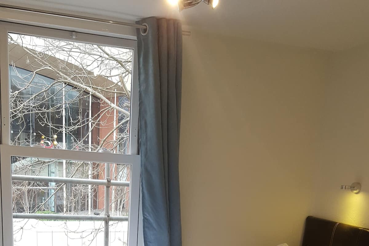 A hotel room with a bed, a window with curtains, a headboard, and a wall-mounted light fixture.