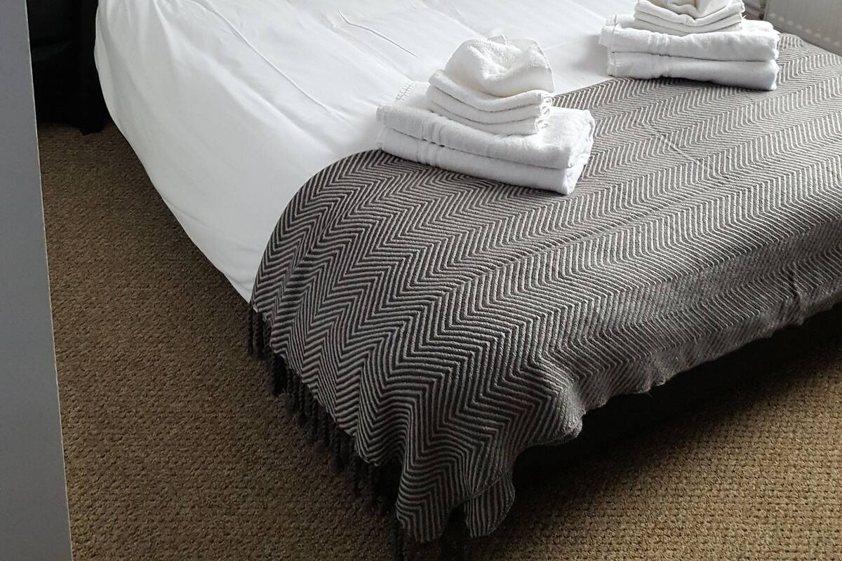 A neatly made bed with white linens and a striped throw blanket, a black nightstand with a lamp, and a power outlet on the wall.