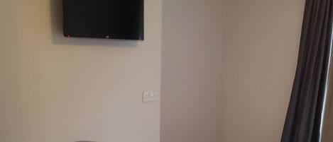 A bedroom with a single bed, a flat-screen TV mounted on the wall, a small alcove, and a dark curtain.