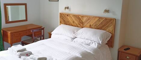 A neatly made bed with white linens and folded towels, a wooden headboard, a bedside table with a remote control, and a small drawer unit.