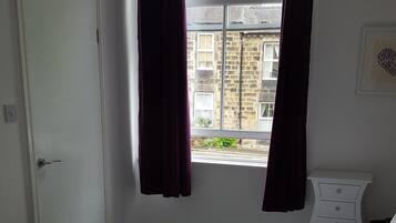 Una cama bien hecha con sábanas blancas, una ventana con cortinas oscuras, un tocador blanco y una puerta a la izquierda.