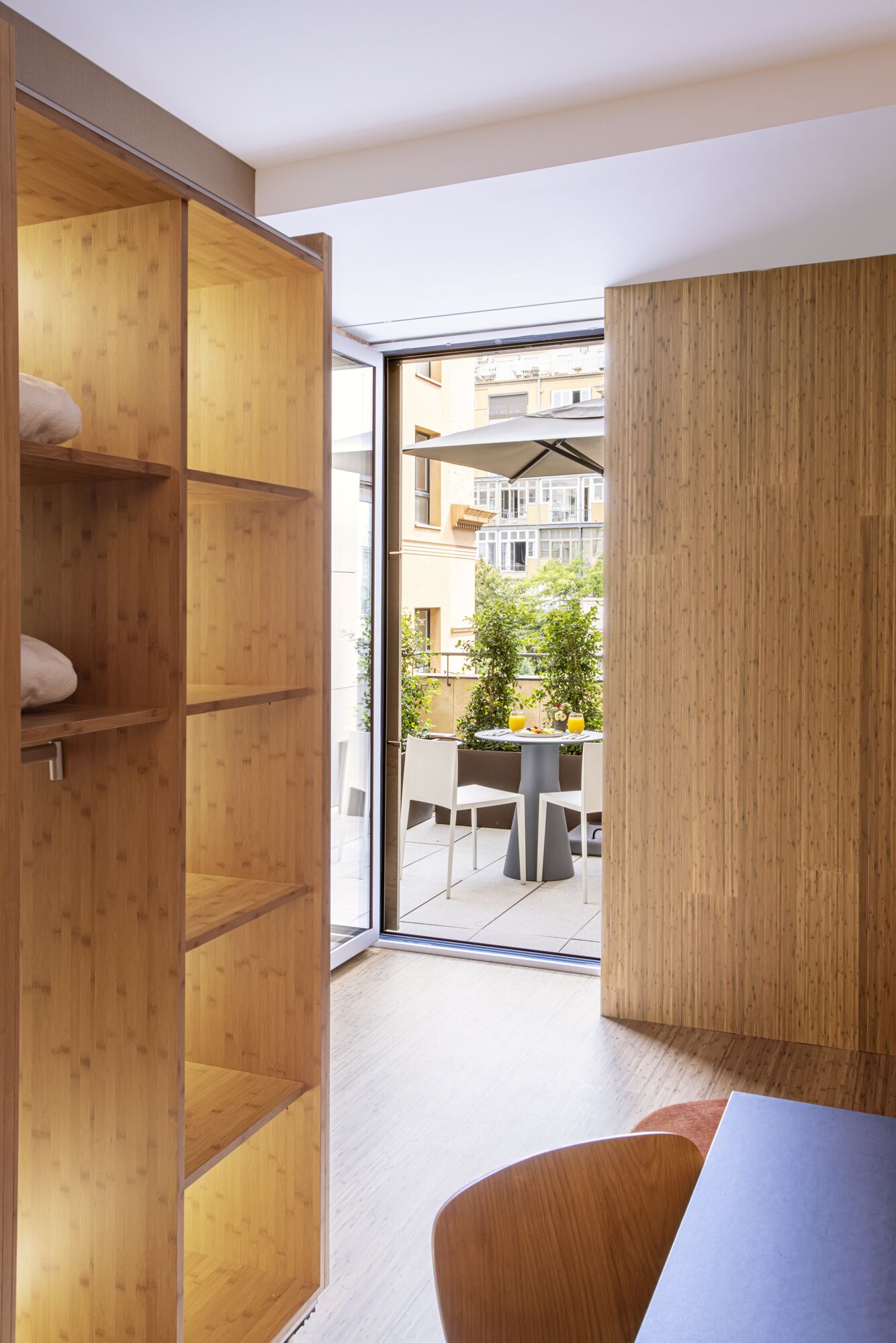 Habitació moderna amb un armari de fusta, una taula de menjador amb cadires i vista a un balcó amb mobiliari exterior i vegetació.