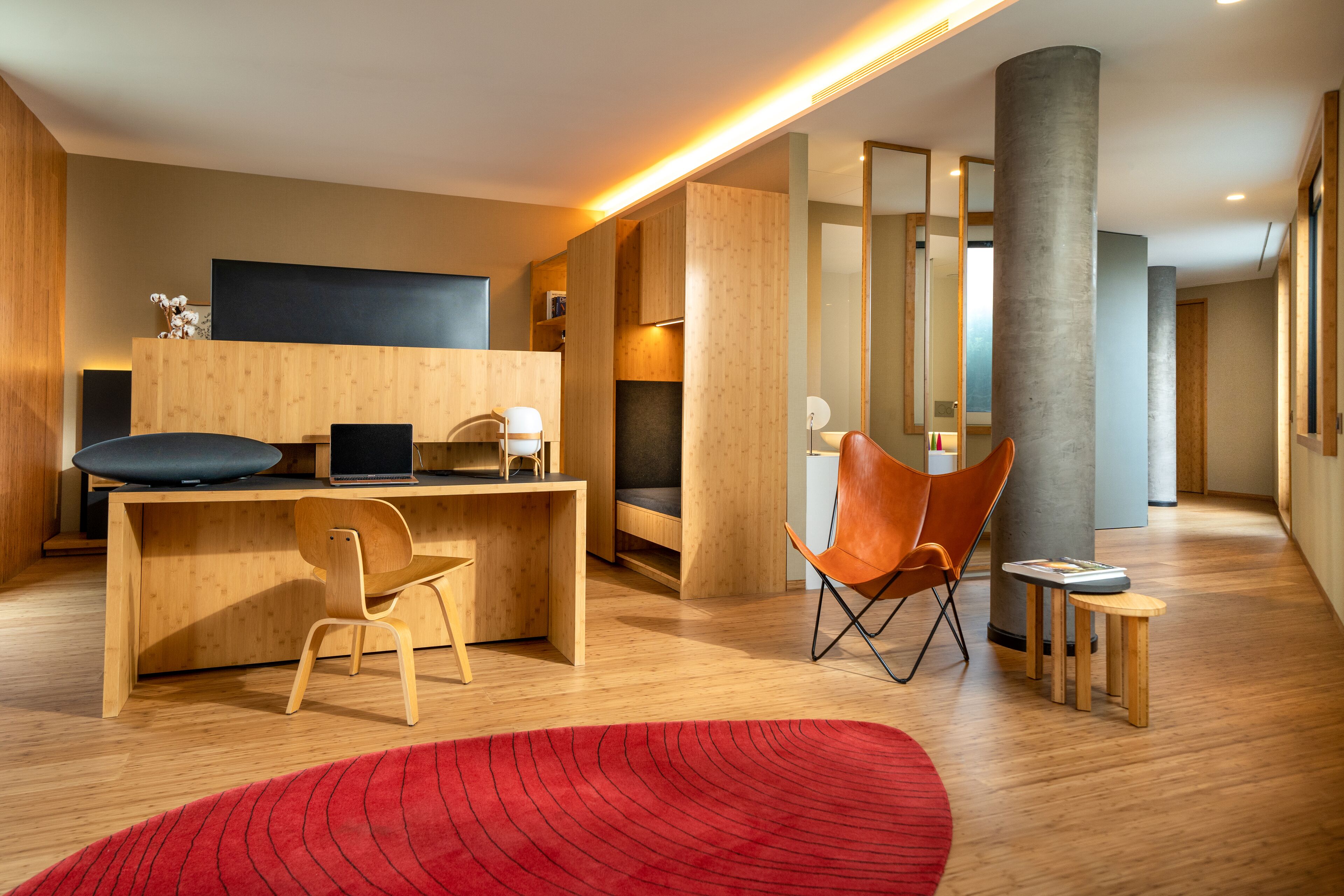 A modern hotel lobby with a wooden reception desk, a large flat-screen TV, a red rug, and a seating area with a wooden chair and a small table.