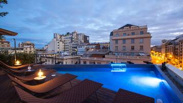 Una piscina en la azotea con sillas de descanso y mesas, rodeada de edificios de la ciudad.