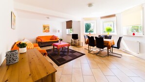 A modern living room with a sofa, a dining table, and chairs. There is a television and a window with a view of greenery. - Landhaus Bettina (Fulda)