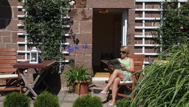 A woman sitting on a bench in a garden area with potted plants and a wooden table. - Landhaus Bettina (Fulda)