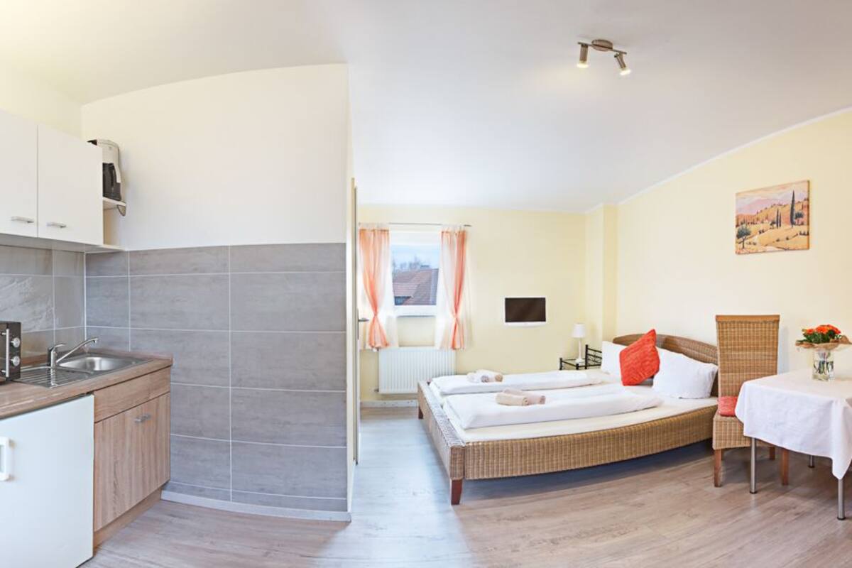 Ein kleines Hotelzimmer mit Küchenzeile, einem Bett mit zwei Kissen, einem Tisch und einem Stuhl.