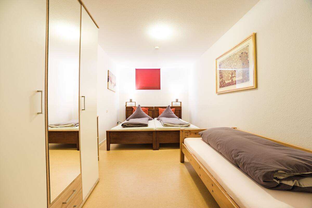 Ein kleines, helles Schlafzimmer mit einem Einzelbett, einem Schreibtisch und einem Stuhl. An einer Seite befindet sich ein Kleiderschrank und an der Wand ein gerahmtes Bild.