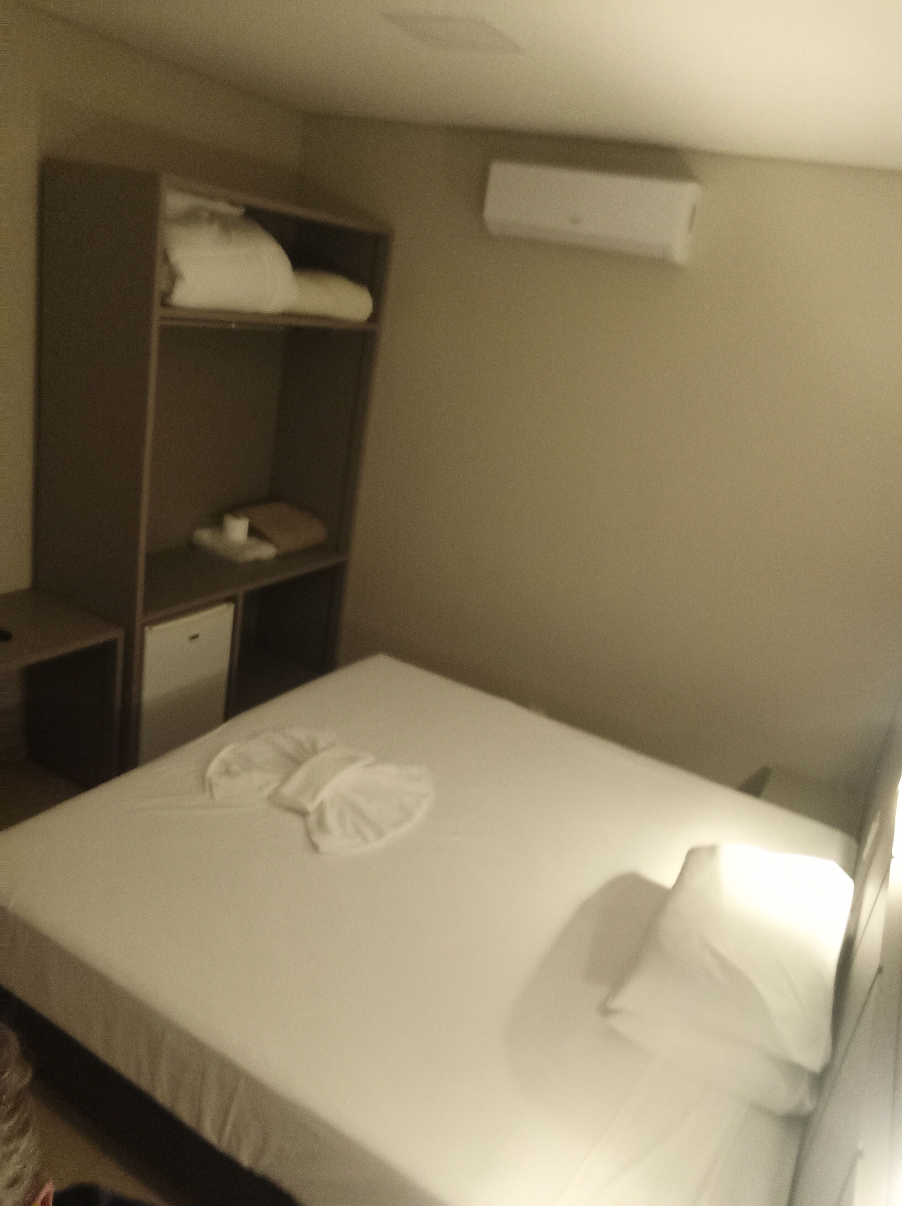 A hotel room with a neatly made bed, a wardrobe, a small desk, and a wall-mounted air conditioning unit.