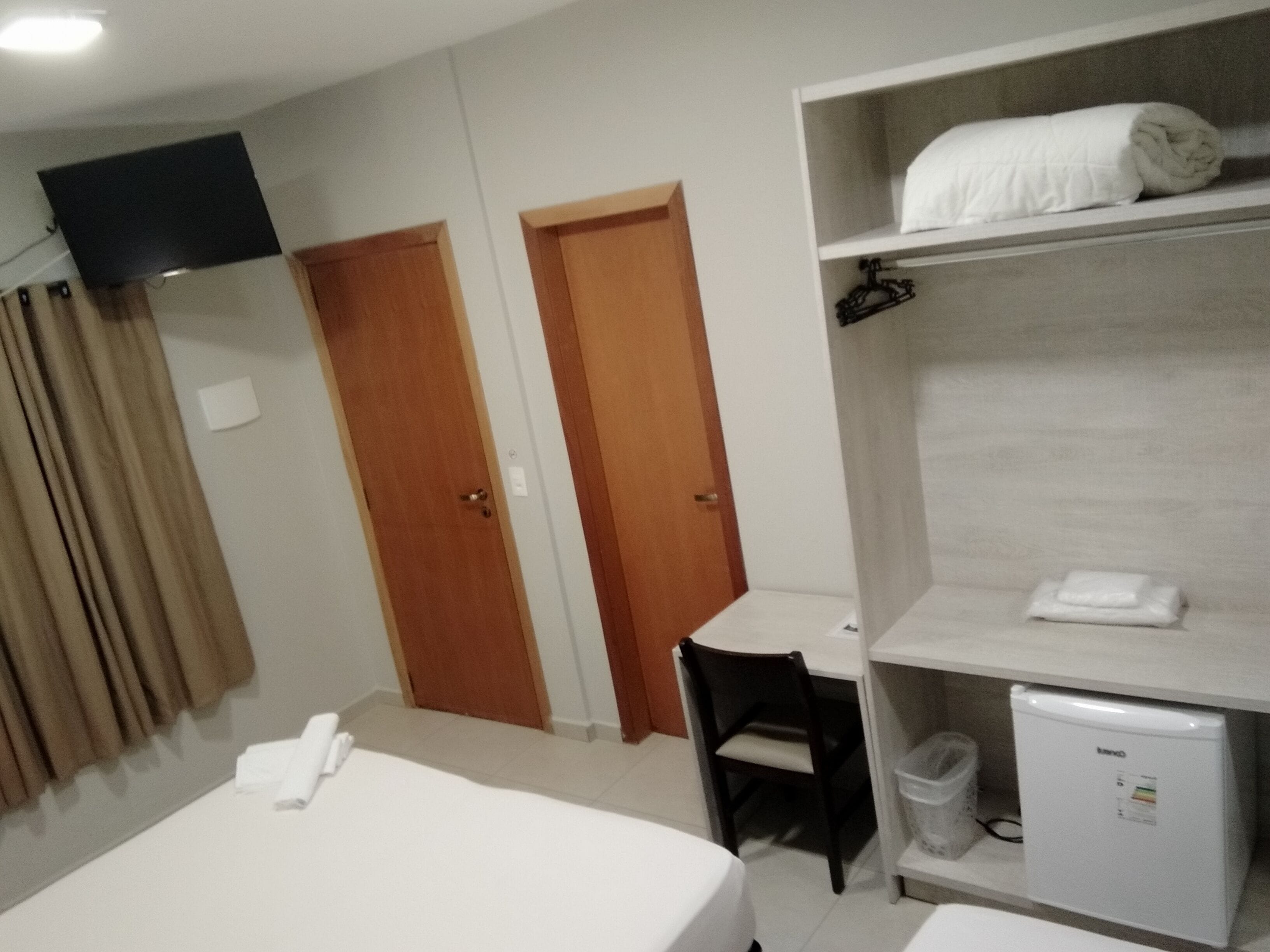 Quarto de hotel com cama, escrivaninha, cadeira, televisão na parede e mini-geladeira.