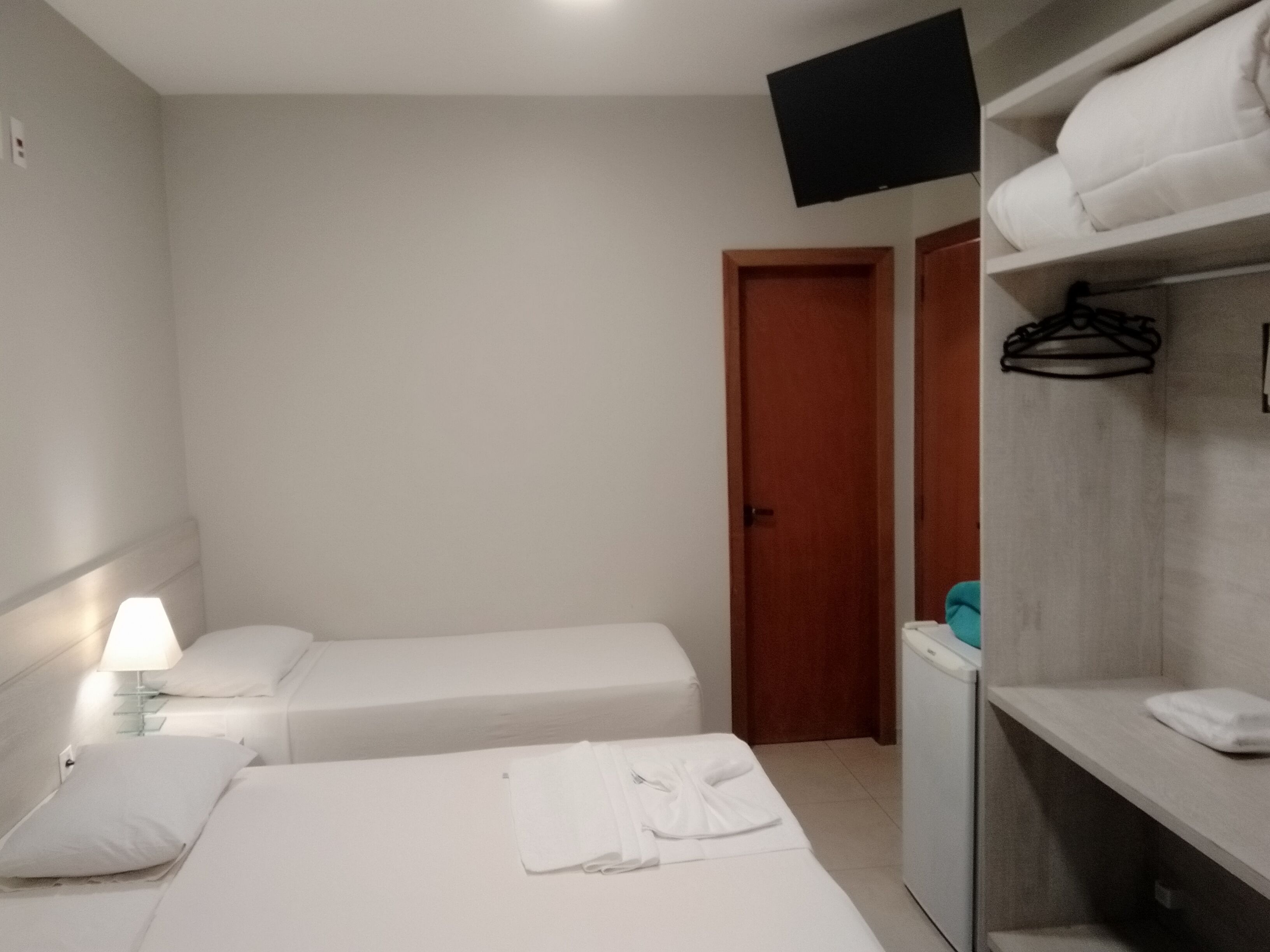 A hotel room with two single beds, a wardrobe, a television mounted on the wall, and a bedside lamp.