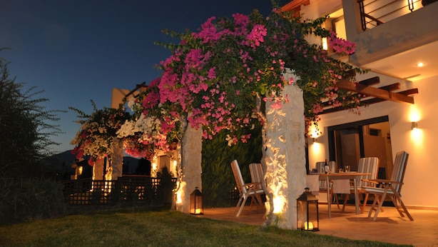 An outdoor patio area with a table and chairs, surrounded by flowering plants and illuminated by wall-mounted lights. - Villa Carlos (Patras)