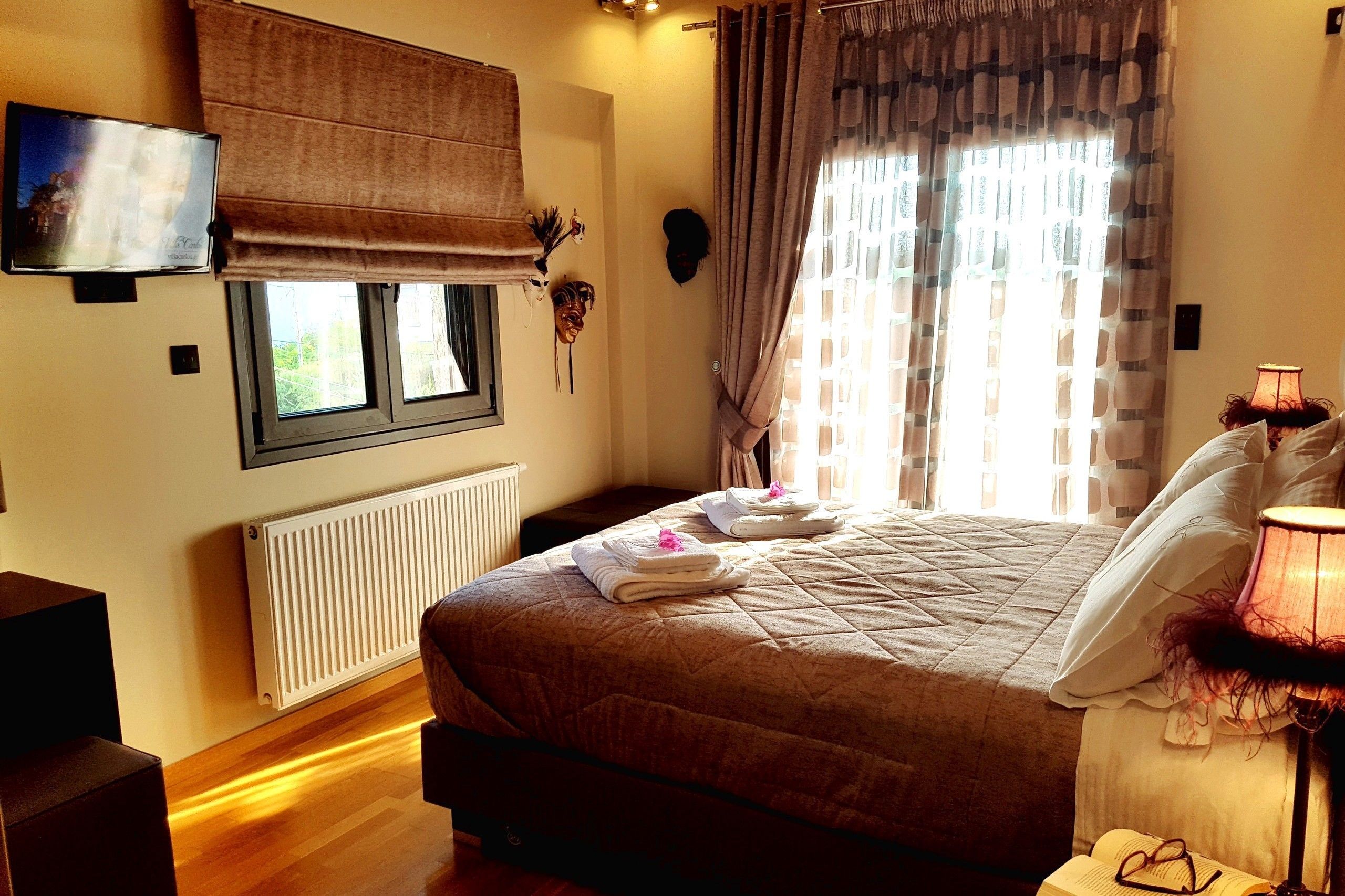 A bedroom with a bed, a television mounted on the wall, a window with curtains, a radiator, and a lamp on the bedside table.