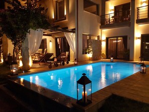 A swimming pool with lounge chairs and a pergola, surrounded by a well-lit patio area with a view of a multi-story building. - Villa Vanessa (Patras)