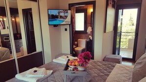 Habitación de hotel con cama, una mesita con frutas y flores, televisor, baño y balcón con vistas.
