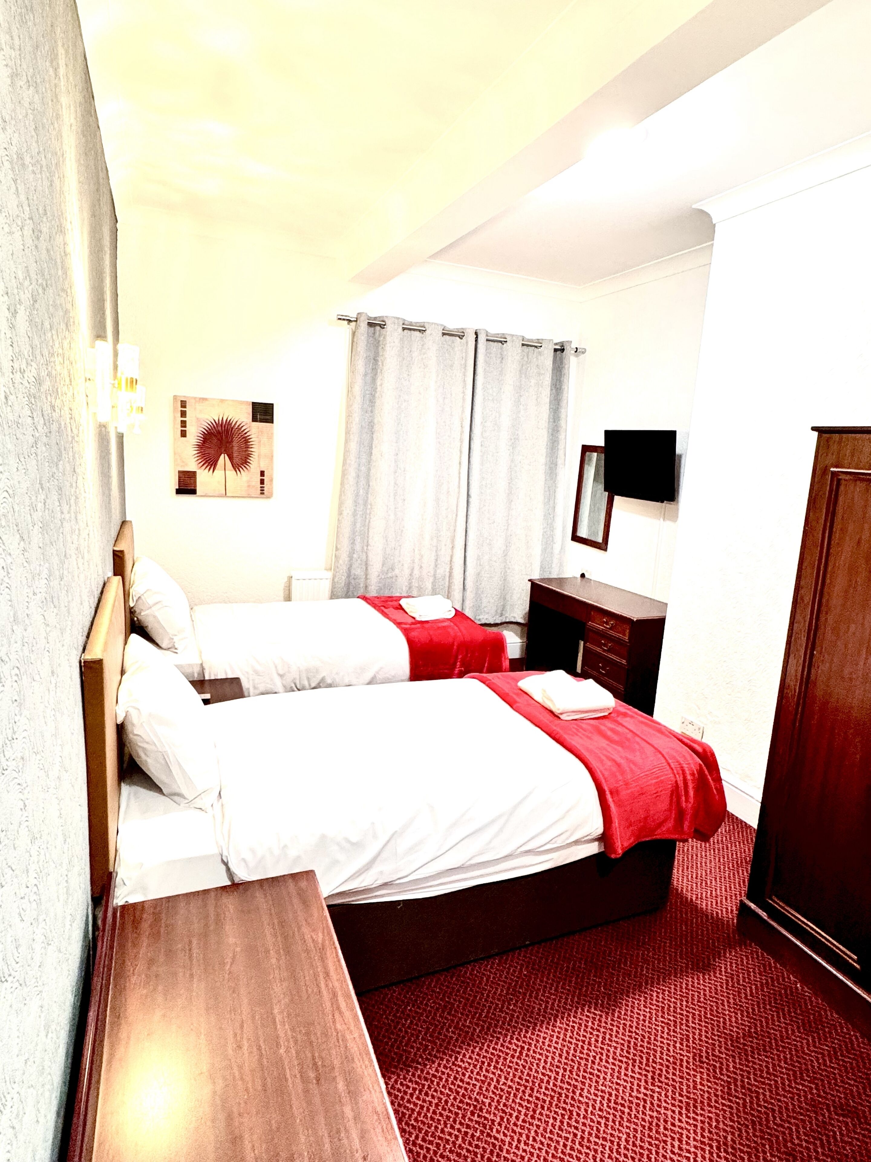 Habitación de hotel con dos camas, una mesita de noche de madera, alfombra roja, televisor montado en la pared y un cuadro.