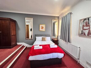 A hotel room with a bed, a wooden wardrobe, a mirror, a painting, and a radiator.