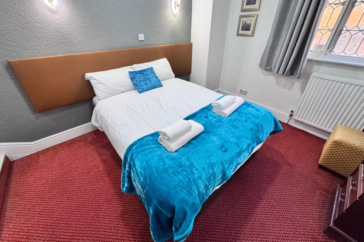 Cama bem arrumada com roupa de cama branca e azul, piso coberto por um tapete vermelho, e uma janela com cortinas.