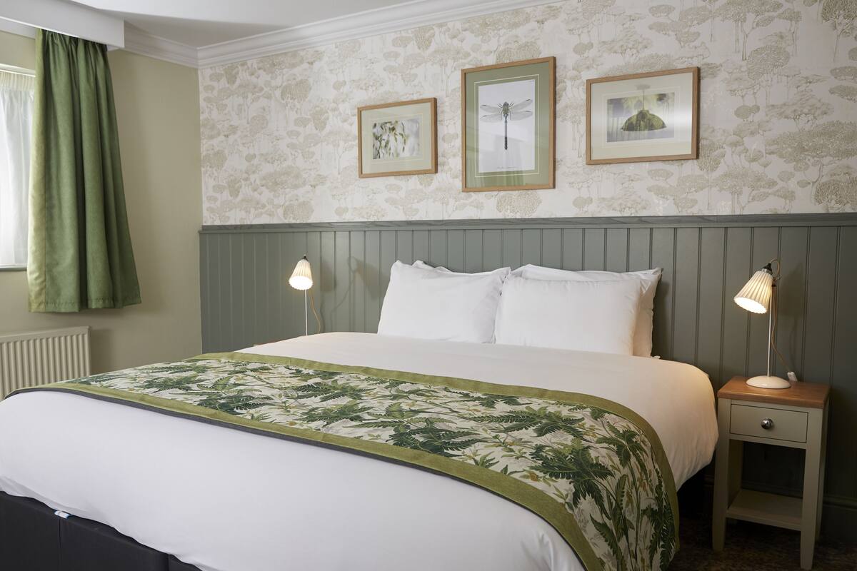 A neatly made bed with a green and white floral bedspread, two bedside lamps, and framed pictures on the wall.