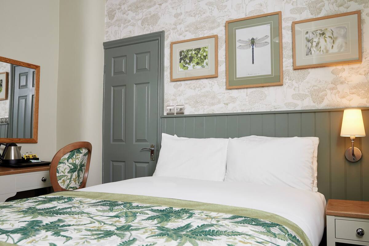 A neatly arranged bedroom with a bed, a dresser, a chair, and a door. There are framed pictures on the wall and a lamp on the nightstand.