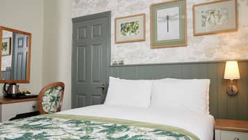 A neatly arranged bedroom with a bed, a dresser, a chair, and a door. There are framed pictures on the wall and a lamp on the nightstand.