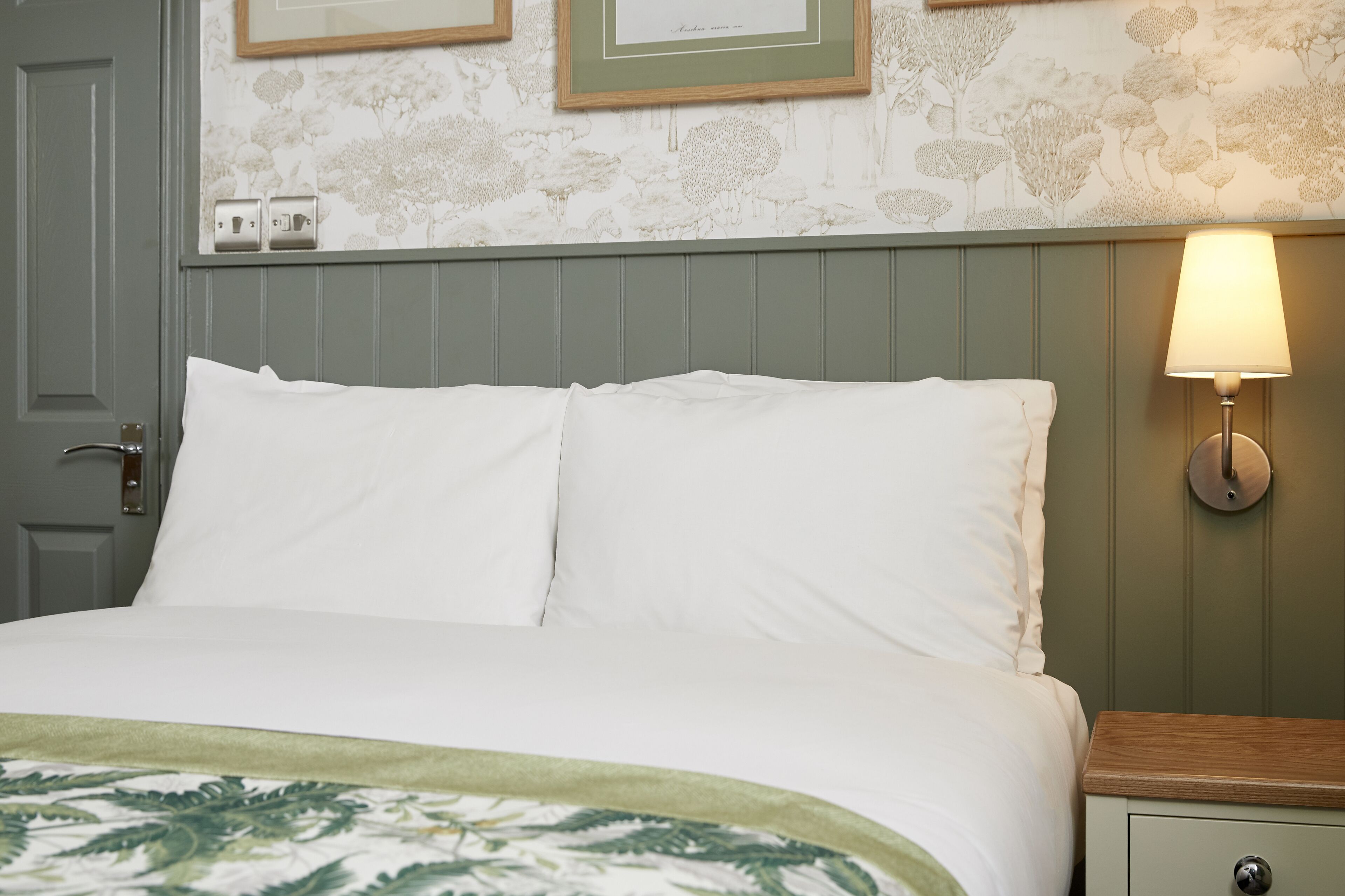 A neatly made bed with white linens, a green throw blanket, and a wall-mounted lamp on the nightstand.