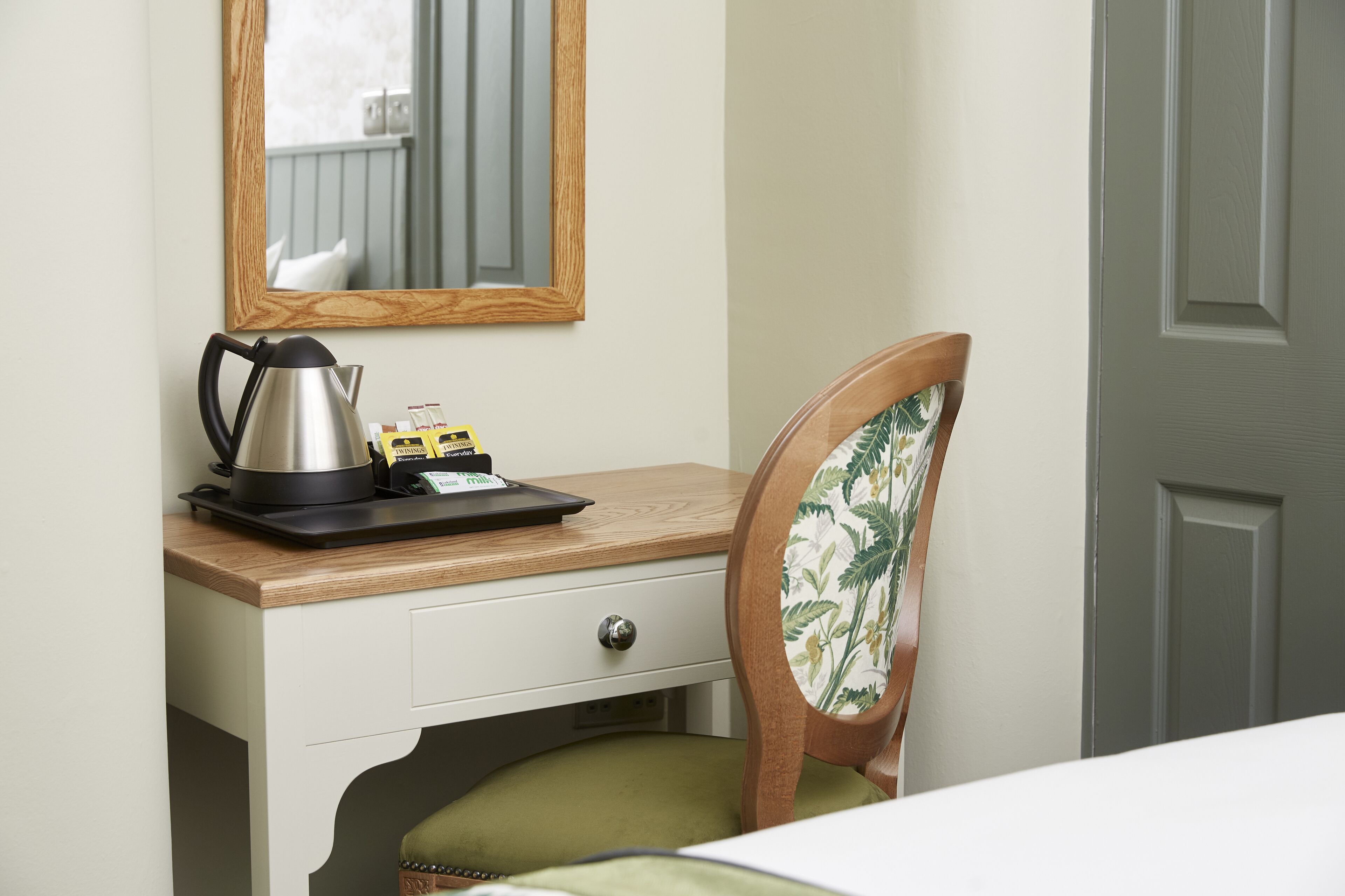 A hotel room with a wooden desk, a chair with floral upholstery, a bed, a door, and a mirror on the wall.