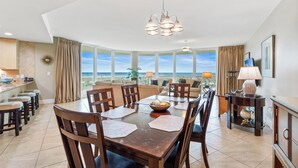 A dining area with a large table, chairs, and a chandelier. There is a view of the ocean through large windows.