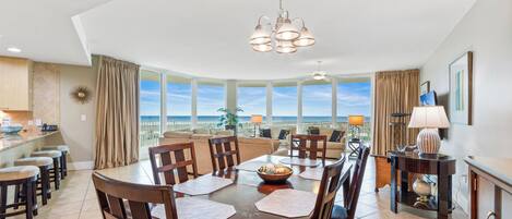A dining area with a large table, chairs, and a chandelier. There is a view of the ocean through large windows.