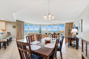 A dining area with a large table, chairs, and a chandelier. There is a view of the ocean through large windows. - Caribe Resort by Hosteeva (Orange Beach)
