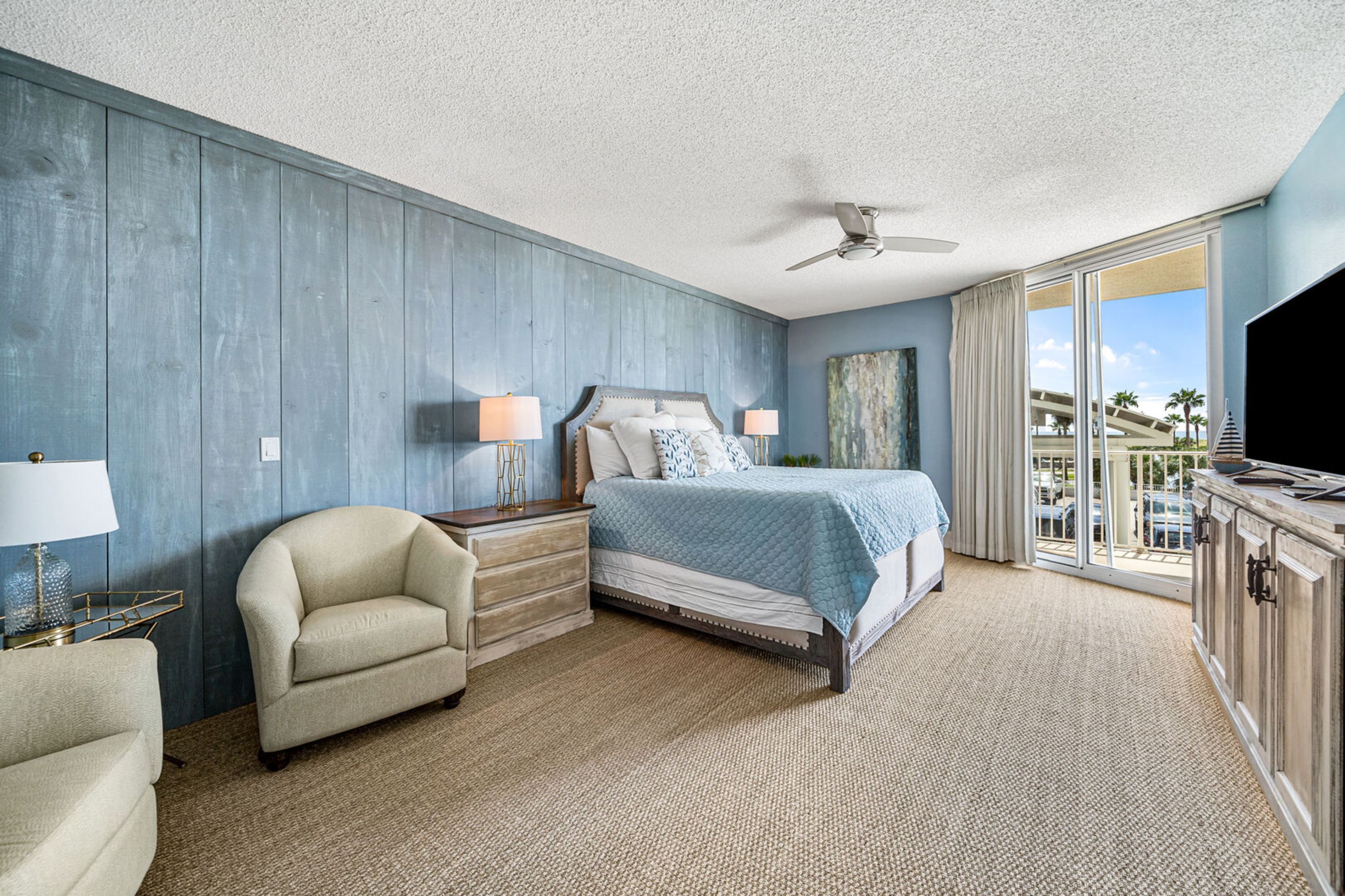 A hotel room with a bed, a chair, a desk with a lamp, a television, and a large window with a view of palm trees and a bridge.