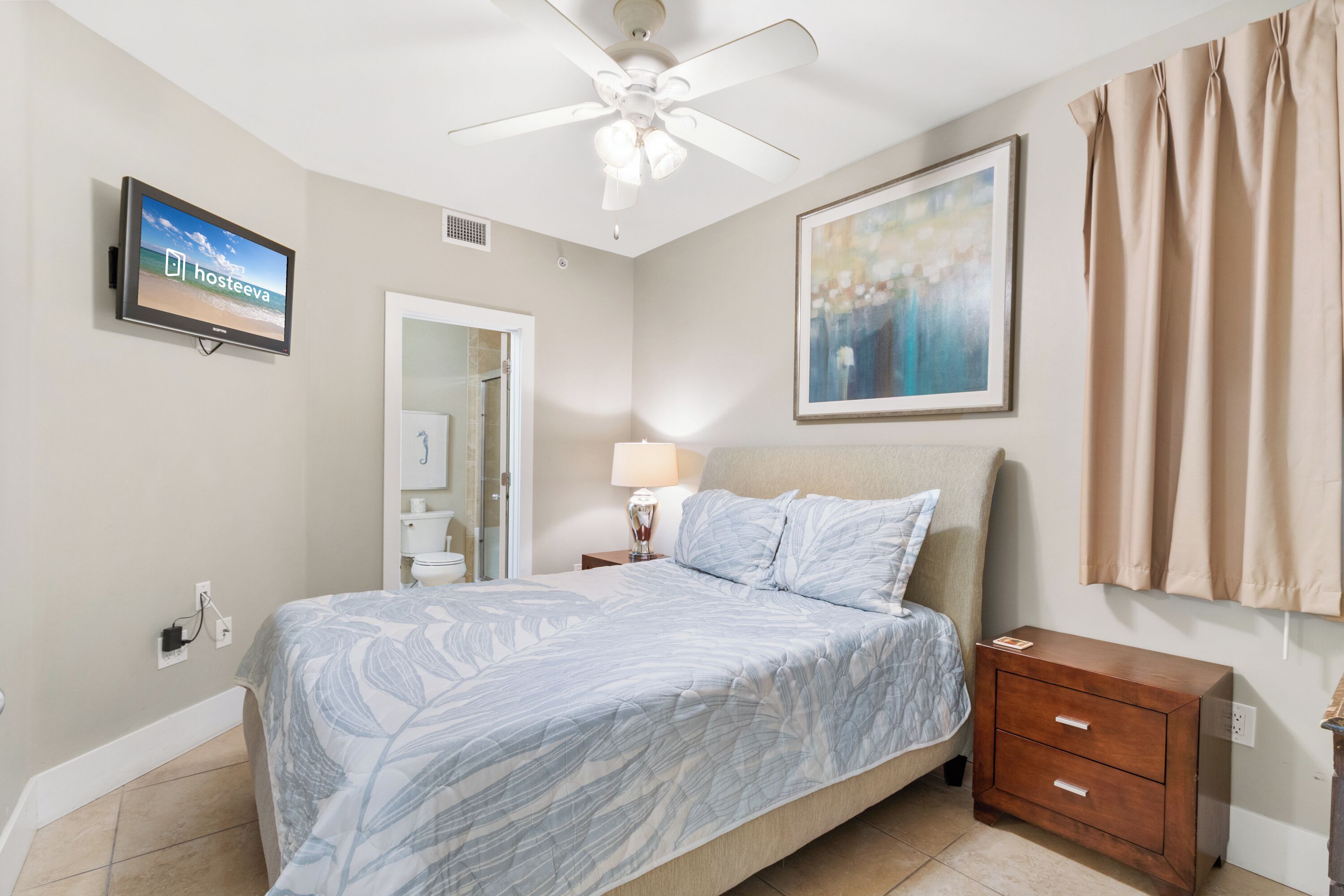 A bedroom with a bed, a nightstand, a TV, a ceiling fan, a painting, and a bathroom door.