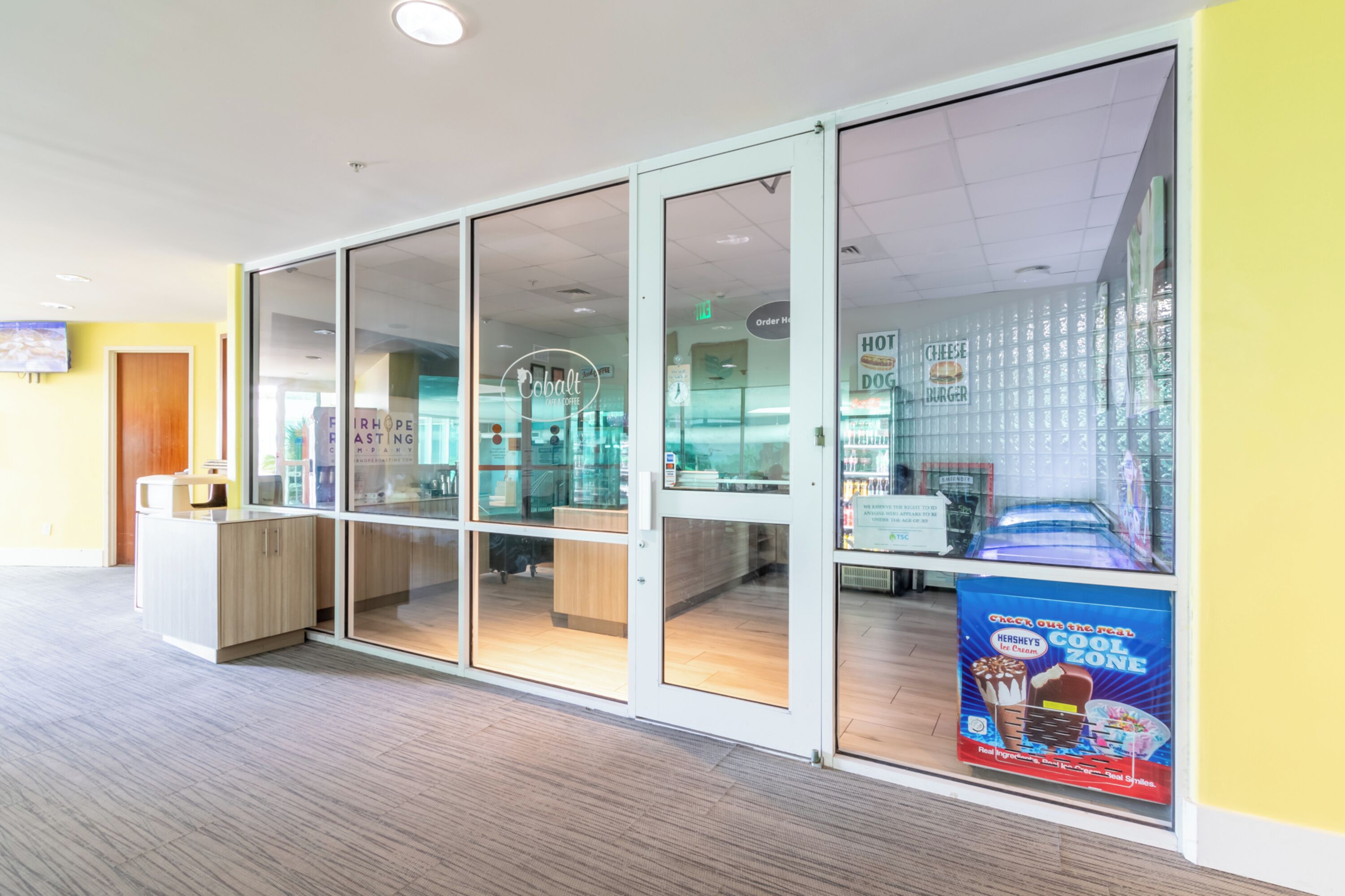 Ein modernes Interieur mit Glastüren, die zu einer Sitzecke, einem Automaten und einem Schild für Hot Dogs und Burger führen.