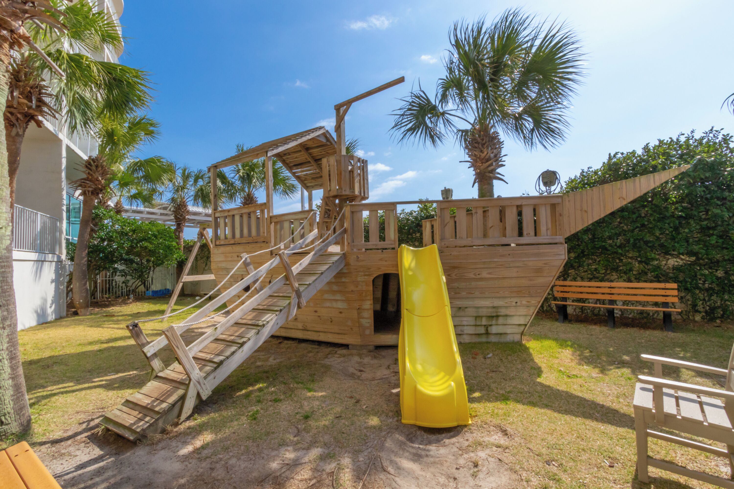 A wooden play structure with a slide and stairs, surrounded by palm trees and a bench.