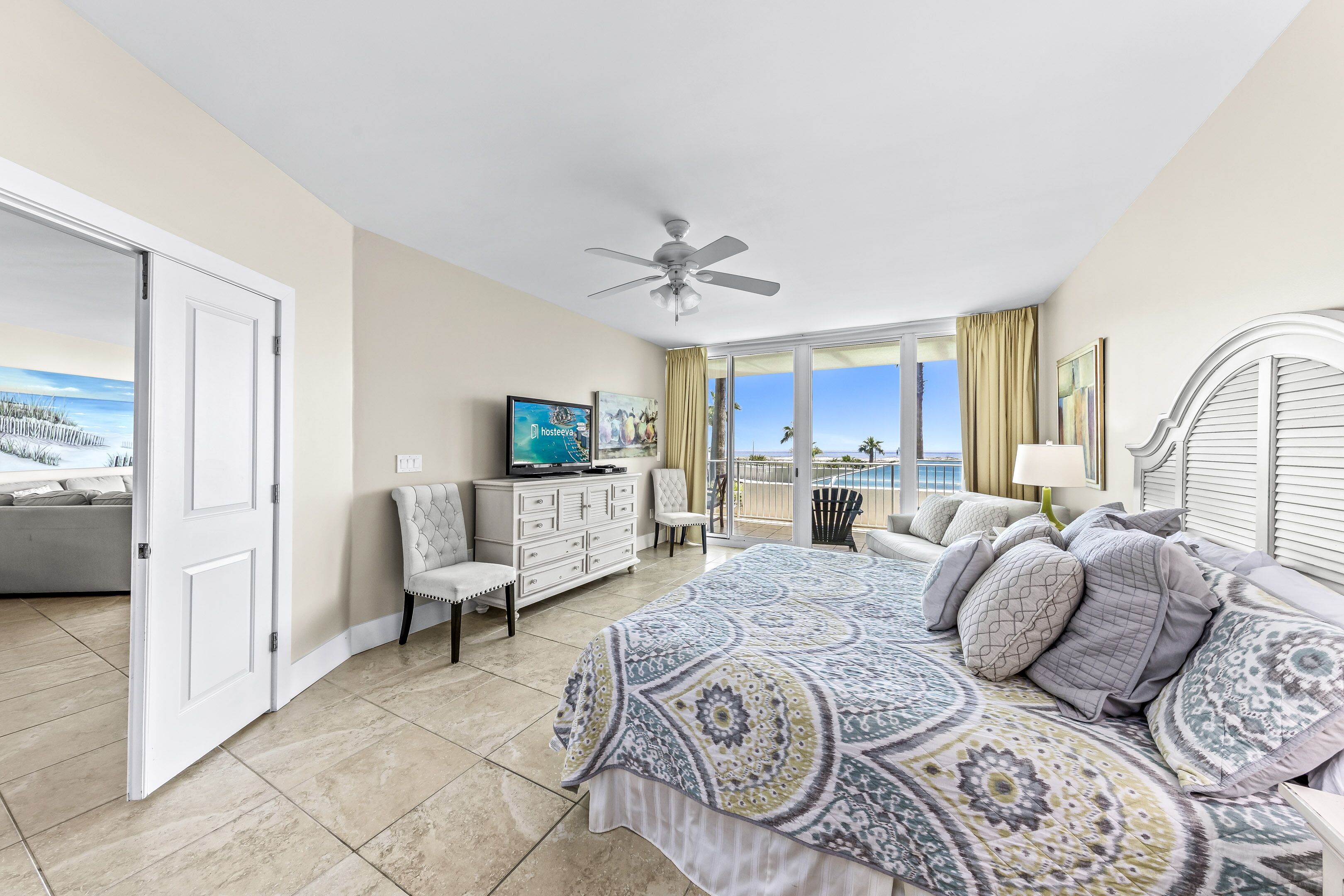 A bedroom with a large bed, a TV, a dresser, and a chair. There is a balcony with a view of the ocean.