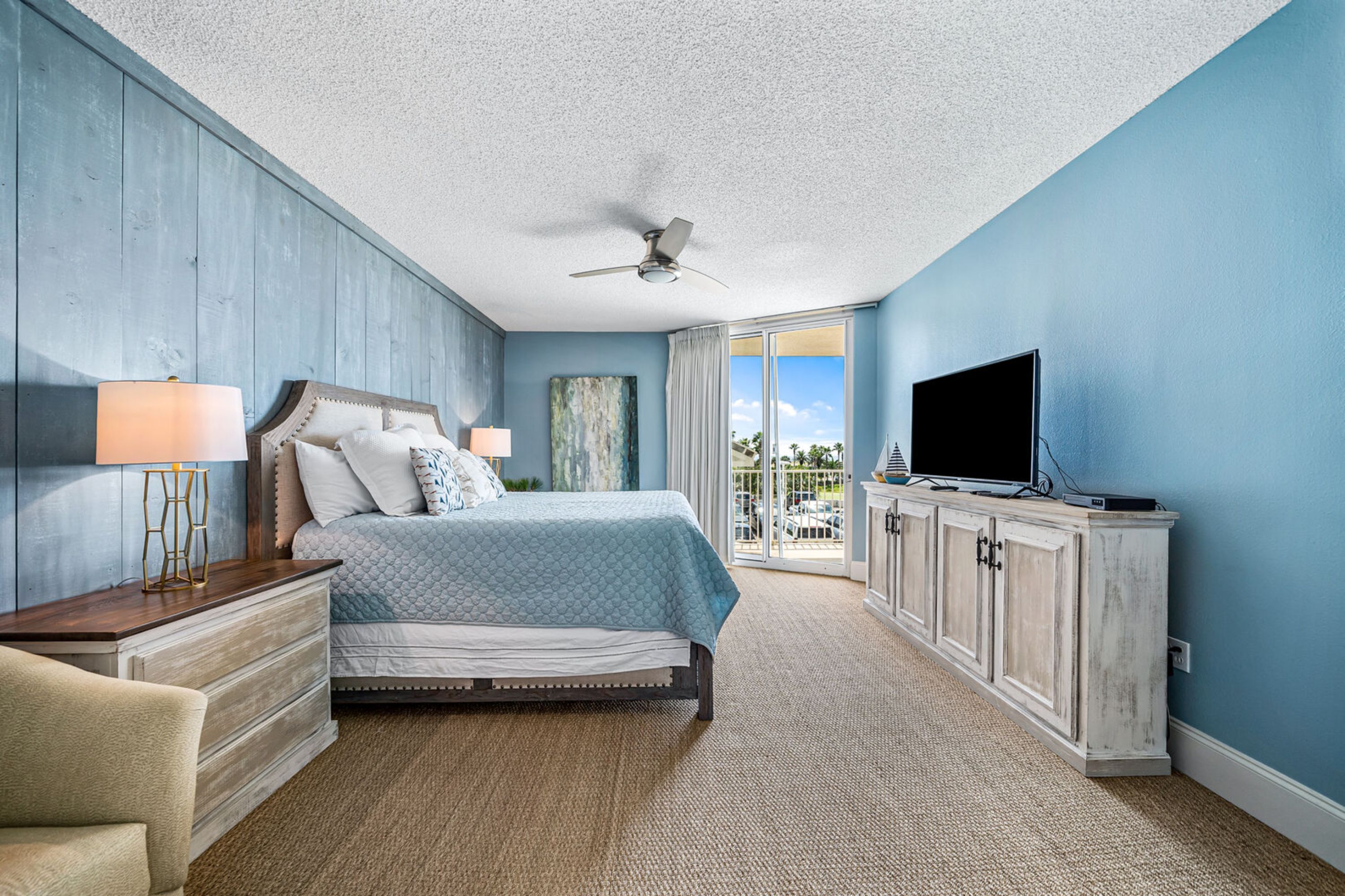 A bedroom with a bed, a TV, a ceiling fan, and a view of the outdoors.
