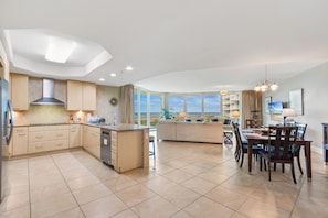 A modern kitchen with a central island, a dining area with a large table and chairs, and a view of the sea through large windows. - Caribe Resort by Hosteeva (Orange Beach)