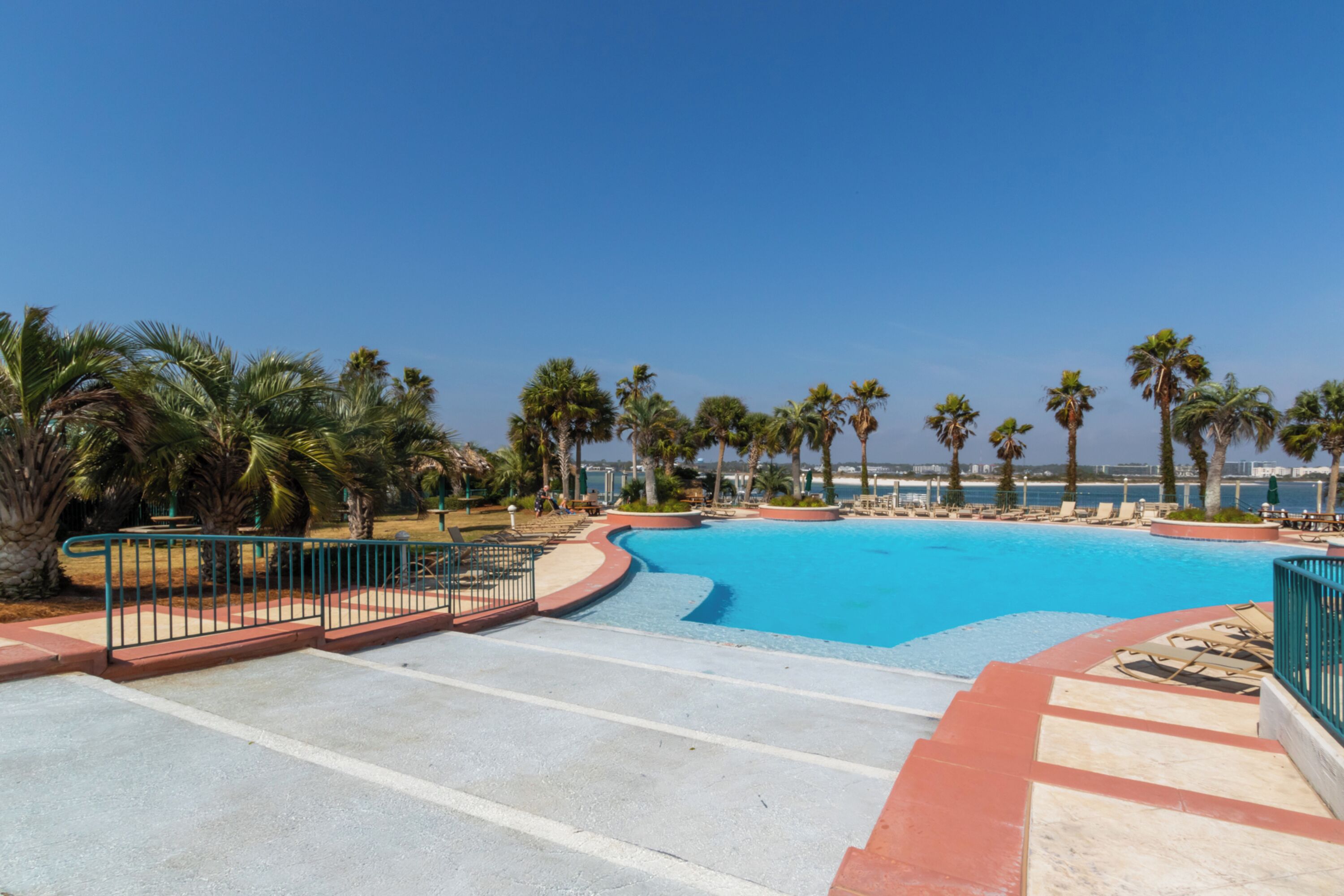 Una amplia piscina exterior con escalones que bajan al agua, rodeada de palmeras y un cielo azul despejado.
