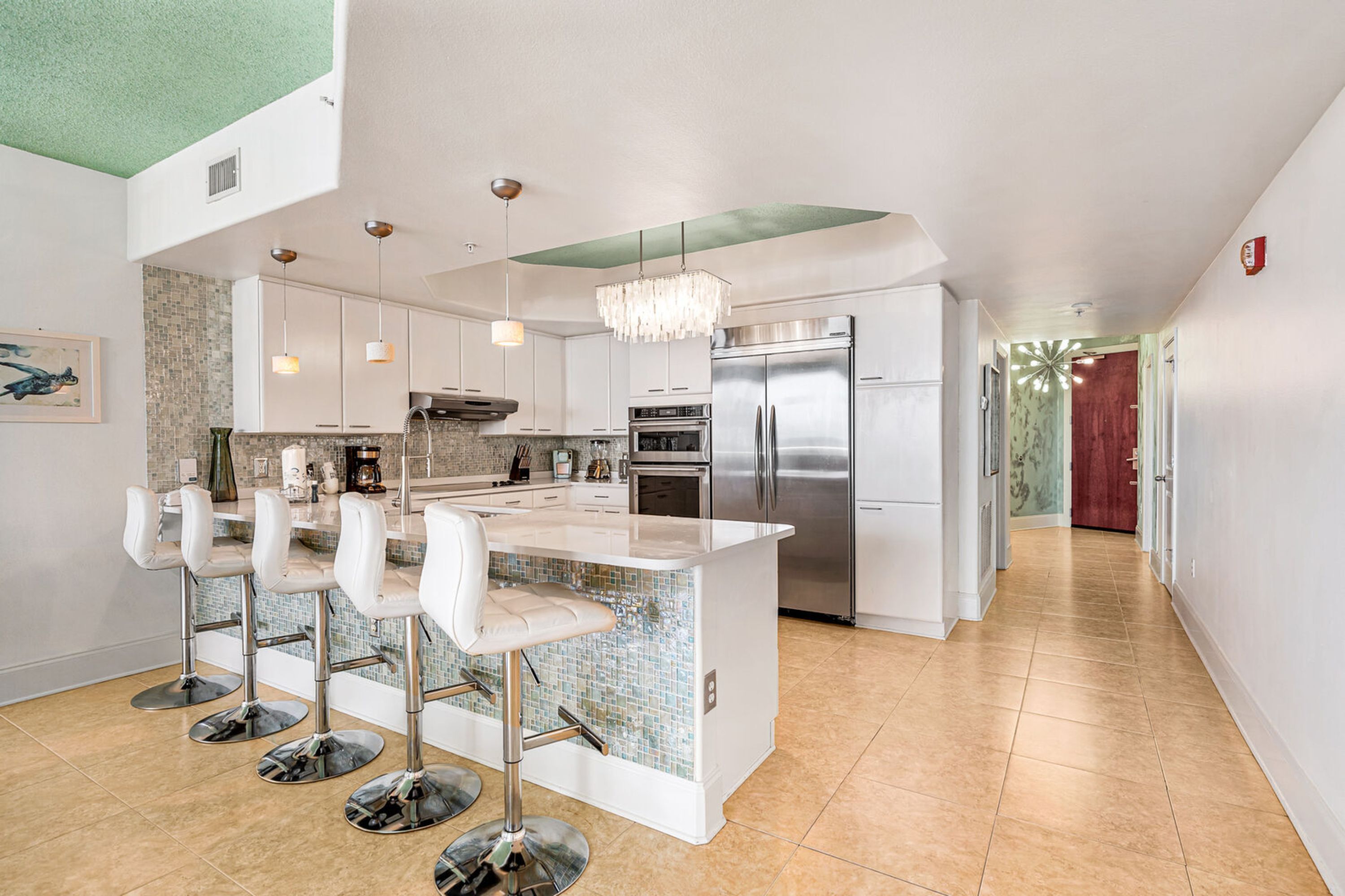 A modern kitchen with a central island, bar stools, stainless steel appliances, and a chandelier.