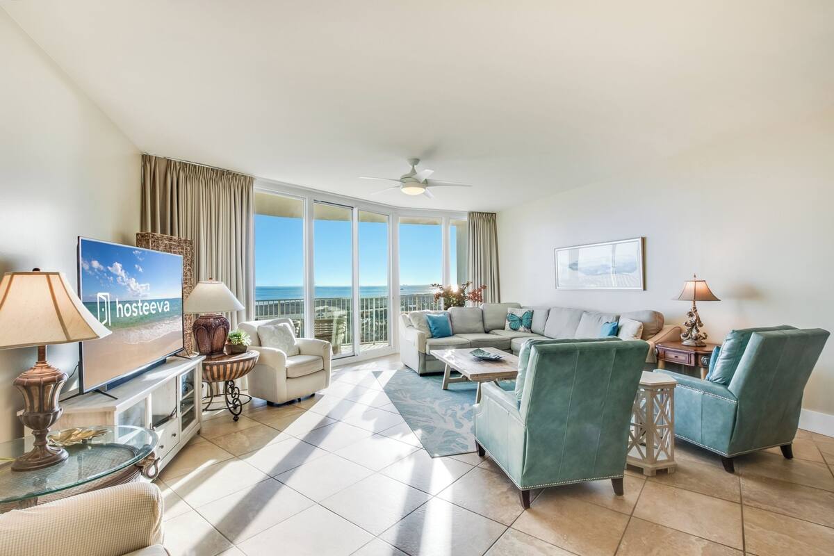 A spacious living room with a large TV, comfortable seating, a balcony with a view, and a ceiling fan.