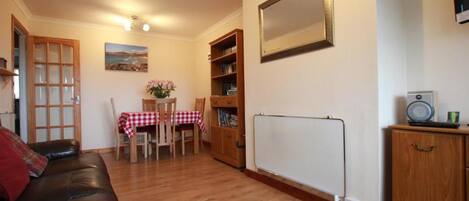 A living room with a leather sofa, a dining table with chairs, a bookshelf, a whiteboard, and a radiator.