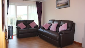 A living room with two brown leather sofas, each with a pink pillow, a wooden floor, a window with white blinds, and a framed picture on the wall. - An Airigh (Stornoway)